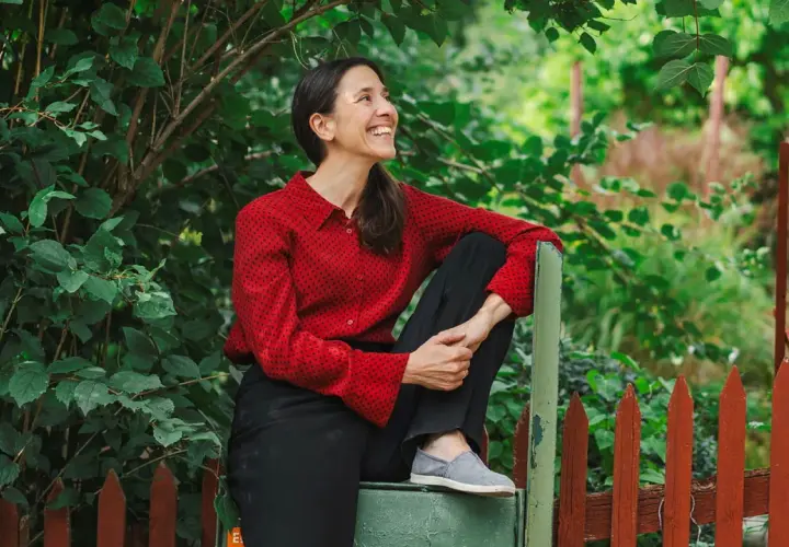 Paloma Grandin sitter avslappnat vid ett staket, omgiven av grönska. Hon ler och tittar uppåt, fångad i ett ögonblick av glädje.
