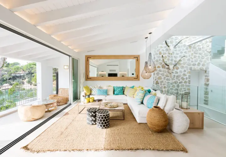 Helles Wohnzimmer mit Finca Style Einrichtung, weißem Sofa, bunten Kissen, Jute-Teppich und Blick auf das Meer.