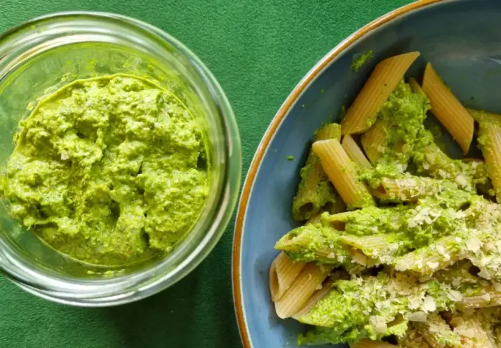 Pesto z liści rzodkiewki w słoiku obok talerza penne z tym zielonym sosem i startym parmezanem.