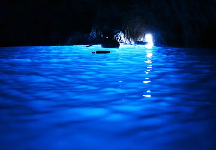 Acqua blu elettrico nella grotta, un piccolo remo galleggia mentre una barca si dirige verso la luce all'uscita della blaue grotte.