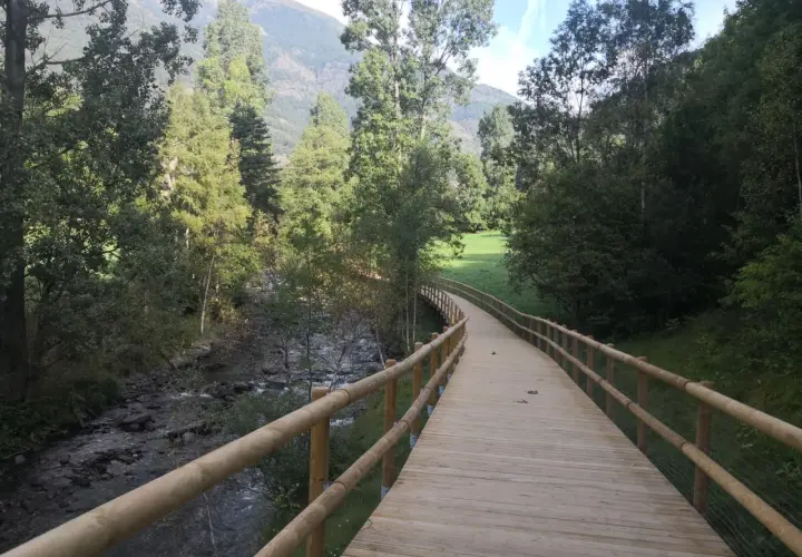 Paseo de madera junto a un río, parte de la ruta del ferro andorra, rodeado de árboles y montañas.