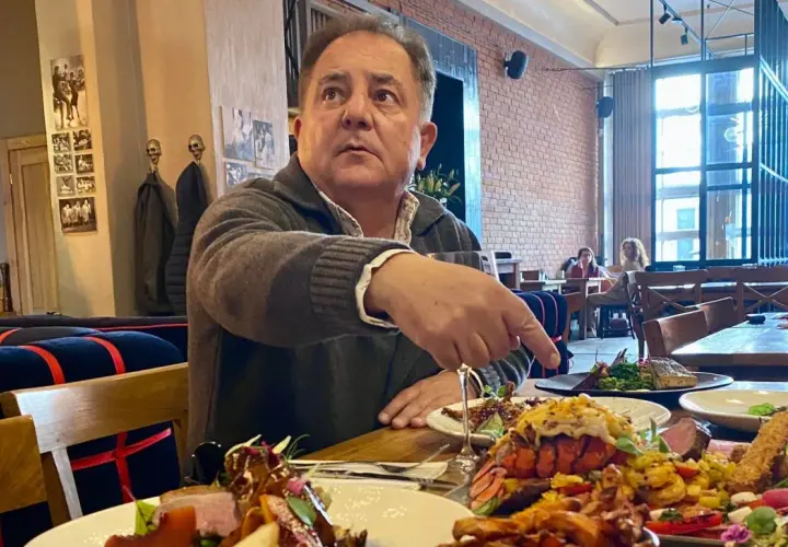 Robert Makłowicz wskazuje na obficie zastawiony stół w restauracji. Widać dania z owocami morza i mięsem.