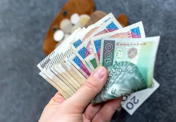 Dłoń trzymająca wachlarz polskich banknotów, w tle portfel z monetami.