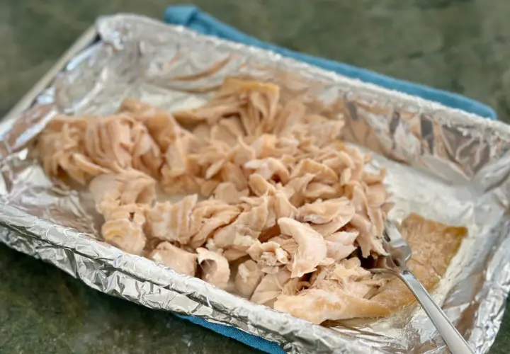 Flaked cooked salmon in a foil-lined baking dish, ready for a salmon flakes recipe. A fork rests on the side.