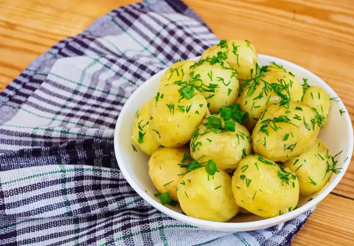 Młode ziemniaki w białej misce, posypane świeżym koperkiem i szczypiorkiem, na tle kratkowanej serwetki i drewnianego stołu.