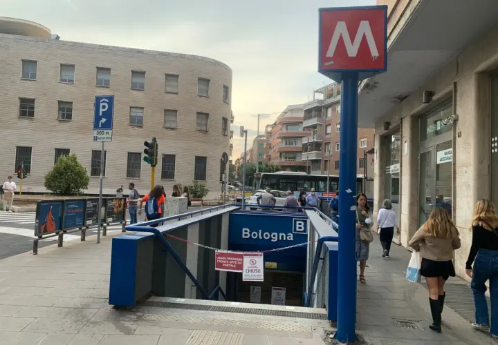 Ingresso della metro Bologna a Roma, con insegna rossa e bianca. Persone camminano sul marciapiede.