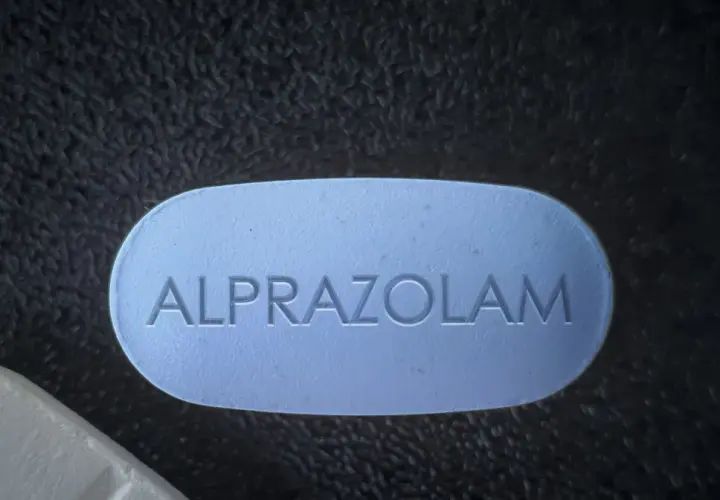 Niebieska tabletka z napisem ALPRAZOLAM leży obok białej tabletki na ciemnym, teksturowanym tle.