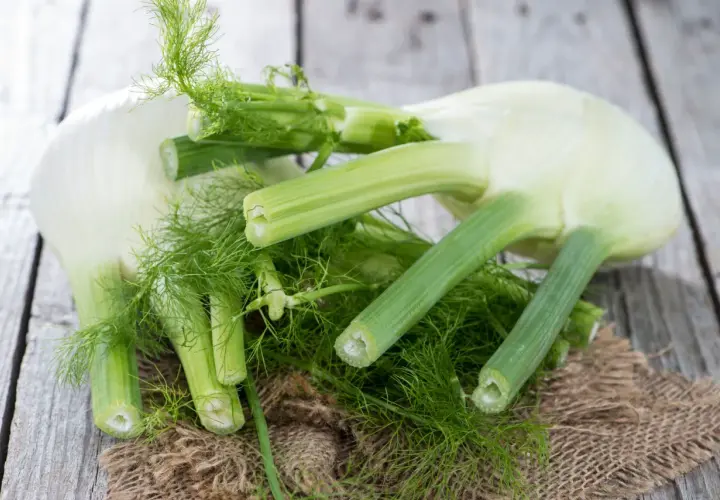 Kilka świeżych bulw fenkułu z zielonymi łodygami i liśćmi, ułożonych na drewnianym blacie. Fenkuł co to? To aromatyczne warzywo.