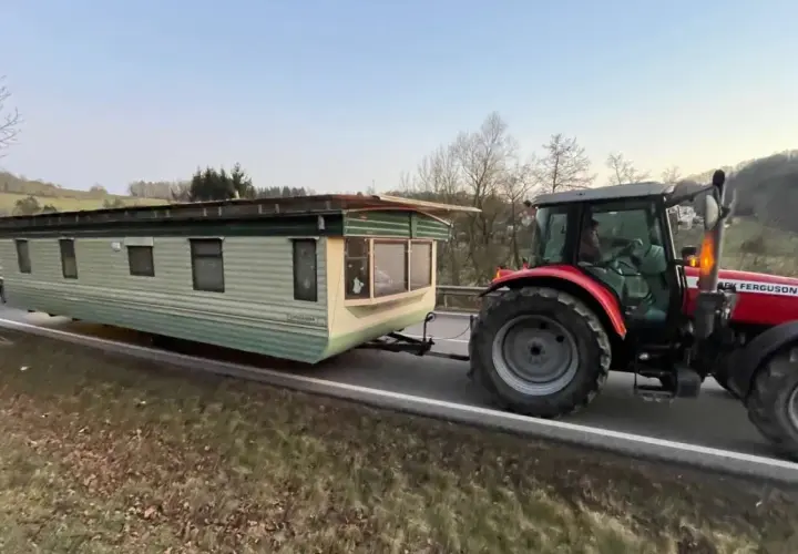 Ein roter Traktor zieht einen Wohnwagen über eine kurvige Straße. Sie sind mit Traktor und Wohnwagen unterwegs, um ein neues Zuhause zu finden.