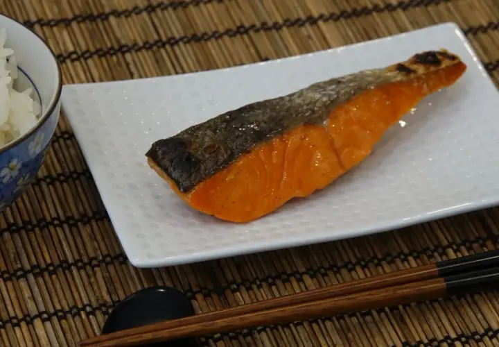 A perfectly grilled salmon shioyaki rests on a white plate, next to a bowl of white rice and chopsticks.
