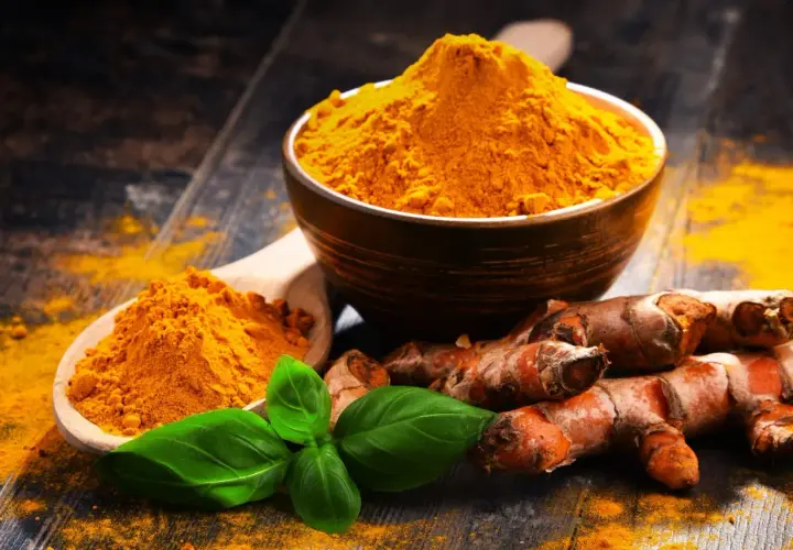 Radici di curcuma e polvere dorata in un cucchiaio e ciotola, con foglie verdi. La curcuma è nota per le sue proprietà antinfiammatorio.