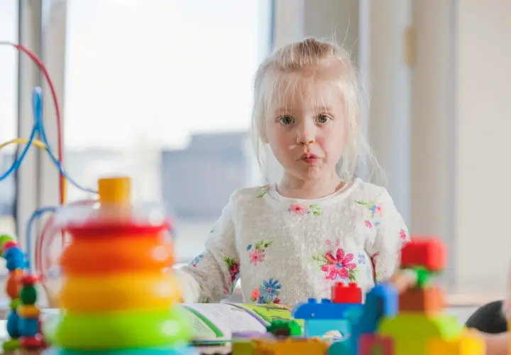 Mała dziewczynka z blond włosami w kwiecistym sweterku bawi się kolorowymi klockami.