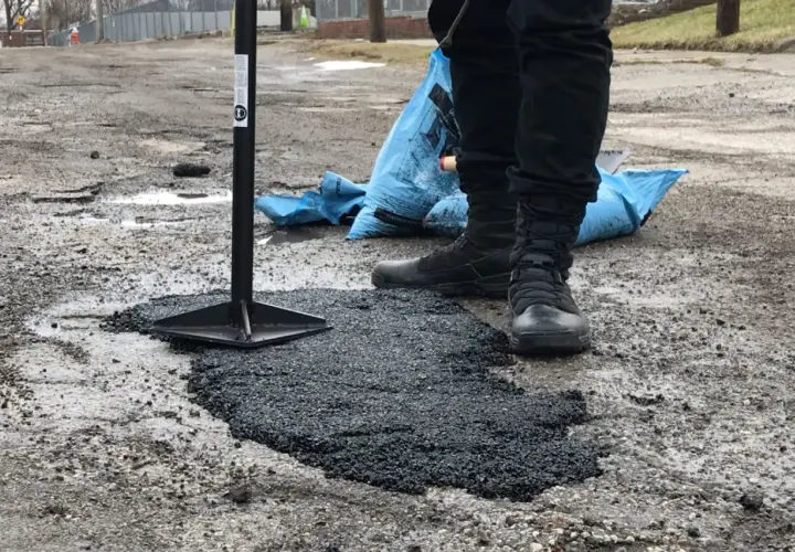 Pracownik ubija asfalt na zimno, naprawiając dziurę w drodze. Obok leży niebieski worek z materiałem.