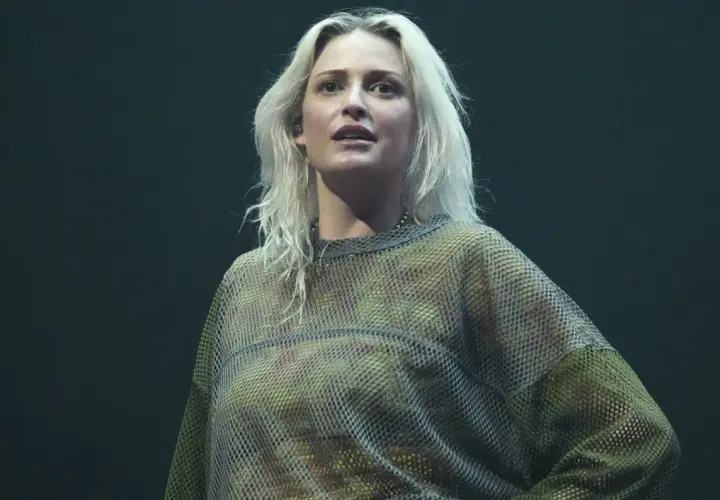 Blond wokalistka w siatkowej bluzce, przypominająca styl wokalisty Linkin Park, na ciemnym tle.