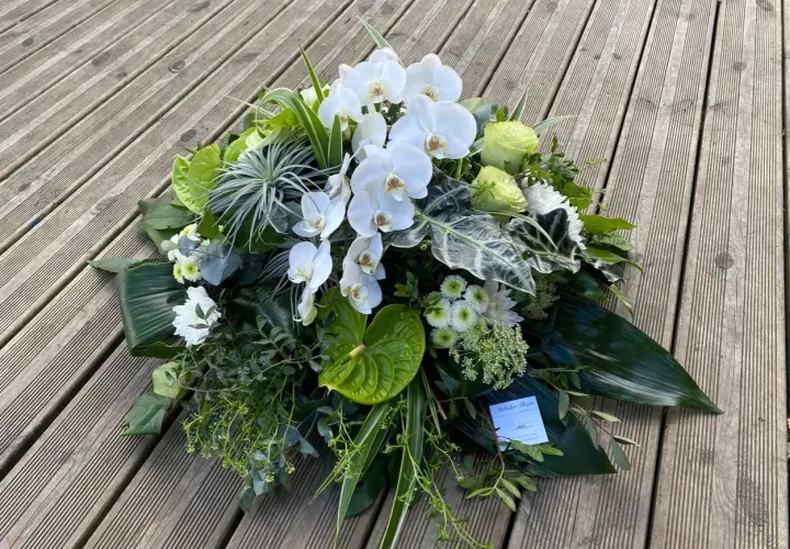 Un arrangement floral blanc et vert, composé d'orchidées, de roses et d'anthuriums, évoque un coussin de deuil moderne.