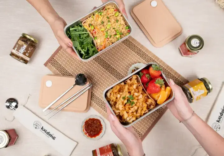 Two hands hold separate noodle bento boxes, one with fried rice and greens, the other with noodles, strawberries, and orange slices.