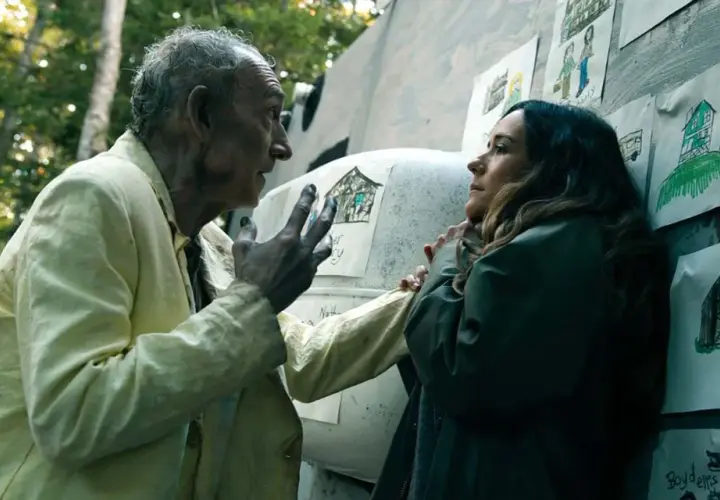 Un vieil homme aux cheveux gris, vêtu d'une chemise jaune, parle à une femme. Des dessins d'enfants ornent le mur derrière eux. Saison 5 date de sortie.