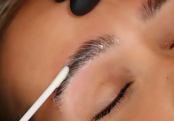 Aplicación de producto para que es el laminado de cejas. Un hisopo blanco aplica una crema sobre las cejas oscuras y peinadas.