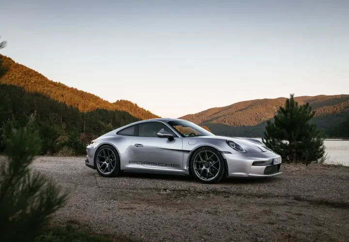 Silberner Porsche 911 ST parkt am Ufer eines Sees, umgeben von bewaldeten Hügeln im warmen Abendlicht.