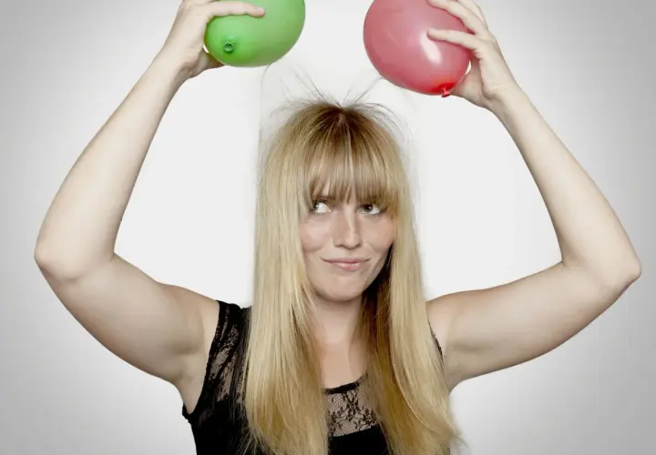En kvinna med blont hår och fräknar håller upp en grön och en röd ballong ovanför huvudet, vilket får hennes hår att resa sig av statiskt elektricitet.