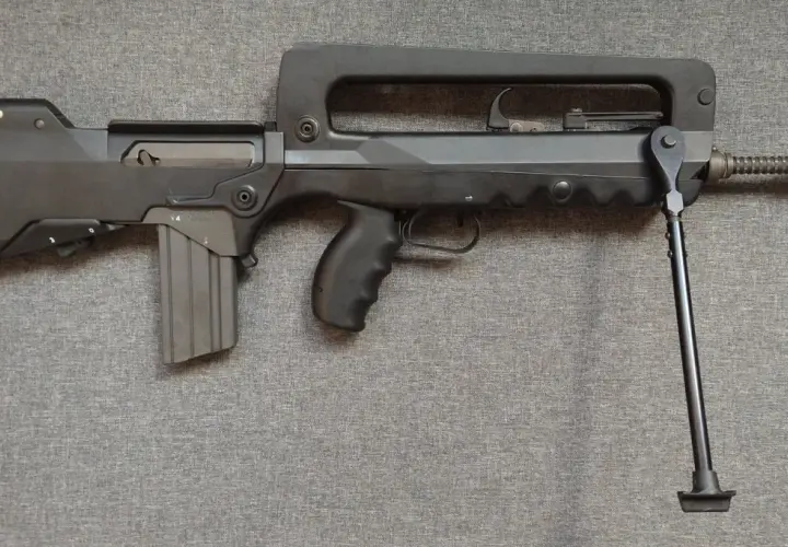Czarny karabin FAMAS z charakterystycznym uchwytem i dwójnogiem, leżący na szarej tkaninie.