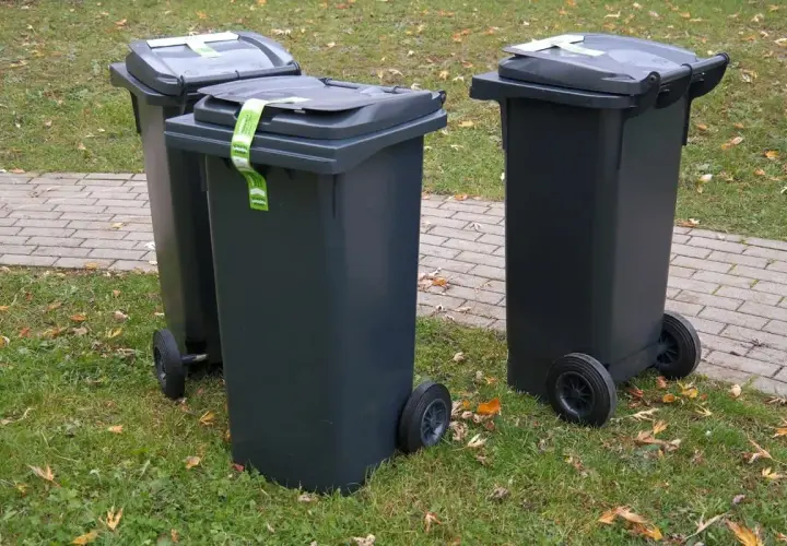 Trzy ciemnoszare kosze na śmieci stoją na trawie obok chodnika. Zastanawiasz się, gdzie wyrzucić piankę poliuretanową? Sprawdź lokalne przepisy dotyczące odpadów.