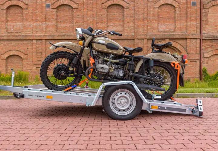 Motocykl Ural na przyczepie z pasami transportowymi, na tle ceglanego budynku.