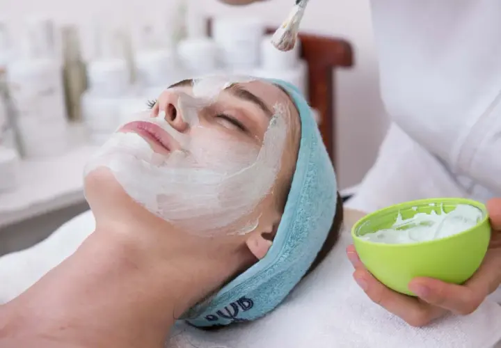 Une femme reçoit un masque ou gommage en premier, appliqué au pinceau sur son visage. Elle est détendue, les yeux fermés.