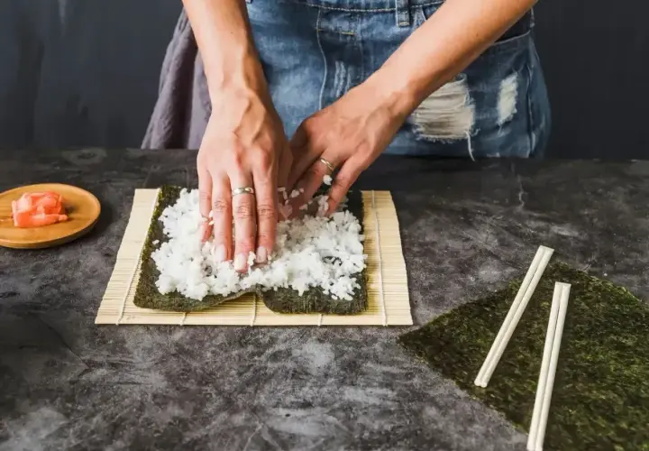 Ręce rozprowadzają ryż na nori, przygotowując zaprawę do sushi. Obok leżą pałeczki i marynowany imbir.