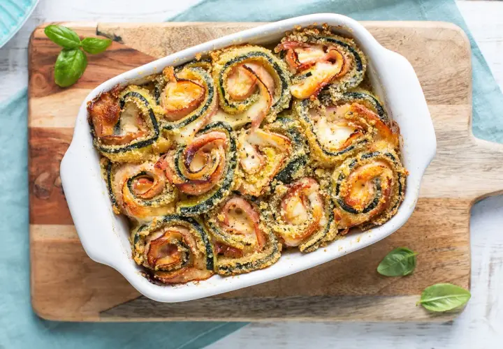 Involtini di zucchine al forno, dorati e filanti, con prosciutto e formaggio, guarniti con basilico fresco.