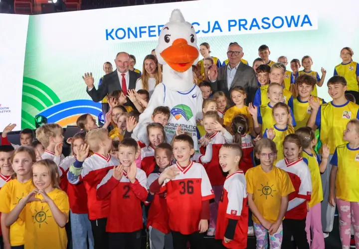 Uśmiechnięte dzieci i duża maskotka z logo lekkoatletycznych mistrzostw świata. Wszyscy pozują do zdjęcia, w tle napis "Konferencja prasowa".