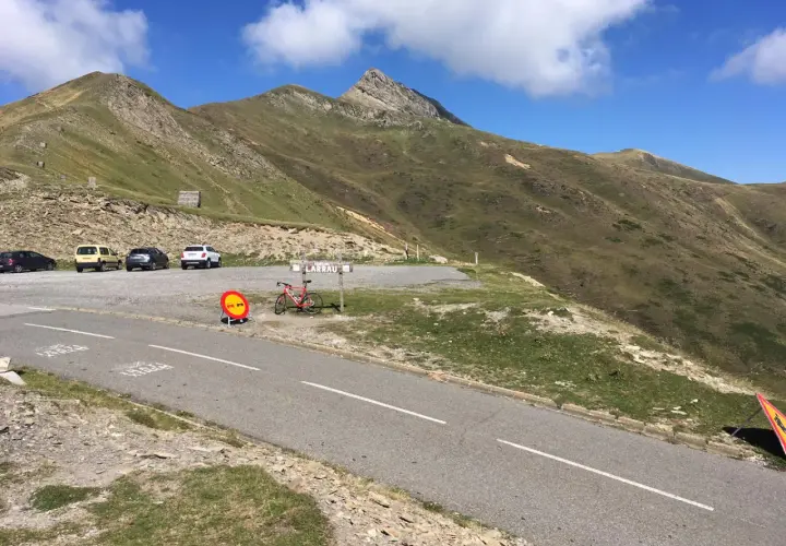 Un ciclista descansa junto a su bicicleta roja en el **puerto de Larrau**, con coches aparcados y montañas verdes bajo un cielo azul.