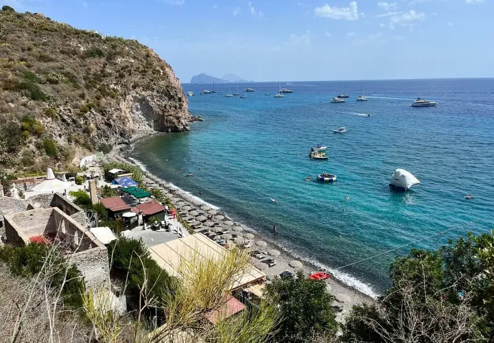 Plage animée à Lipari, avec des parasols, des petits restaurants et des bateaux naviguant sur la mer turquoise.