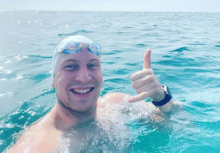 Uśmiechnięty pływak w czepku i goglach, z ręką w geście "hang loose", w trakcie przepłynięcia Bałtyku wpław.