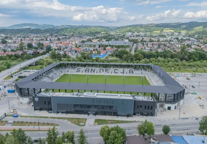 Nowoczesny stadion Sandecji z zieloną murawą i trybunami, otoczony miastem i wzgórzami.