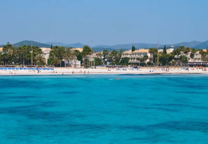 Türkisblaues Meer vor Sa Coma Altstadt mit Palmen und Hotels. Menschen genießen den Strand und das Wasser.