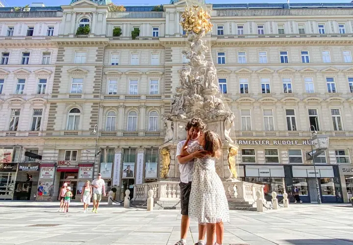 Due bambini si abbracciano davanti alla Colonna della Peste a Vienna. Altri bambini passeggiano sullo sfondo.