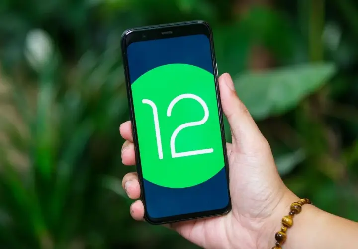 Dłoń trzyma smartfon z logo Android 12 na ekranie, na tle zieleni.