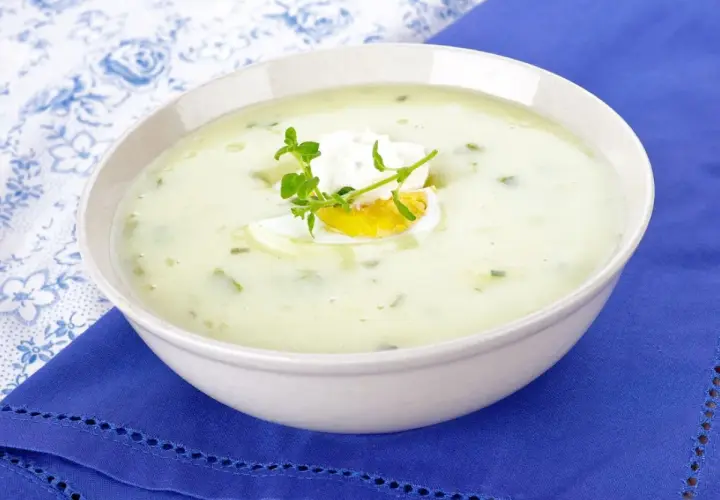 Kremowa zupa z sałaty z jajkiem i ziołami, podana w białej misce na niebieskim obrusie.