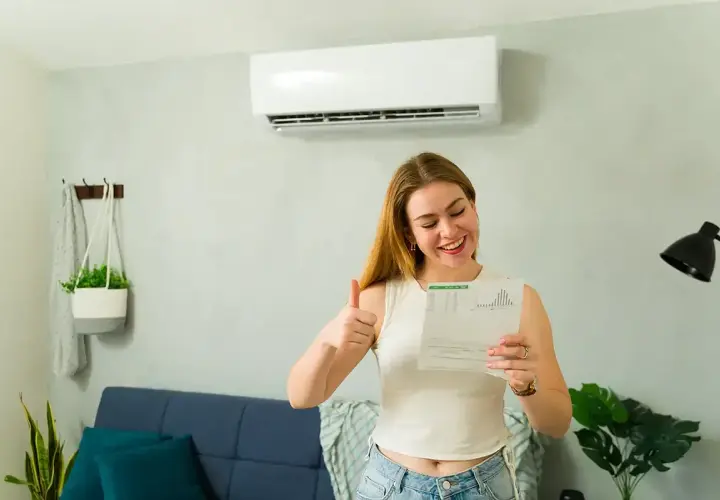 Uśmiechnięta kobieta pokazuje kciuk w górę, trzymając dokumenty. Klimatyzator na ścianie sugeruje, że to może być związane z dofinansowaniem do klimatyzacji.