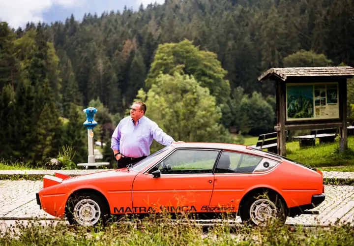 Ein Mann lehnt an seinem leuchtend orangen Simca Matra Bagheera. Im Hintergrund ein dichter Nadelwald.