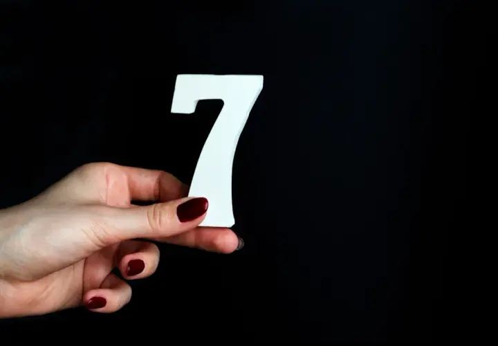 Dłoń z ciemnoczerwonym lakierem trzyma białą cyfrę 7, symbolizującą numerologiczną 7.
