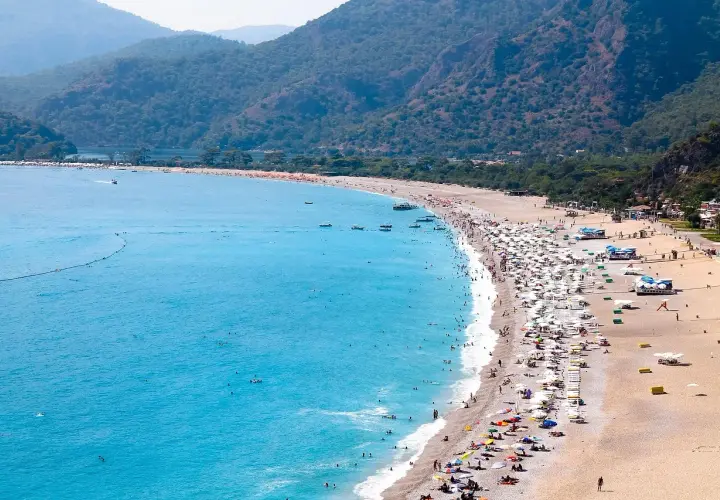 Tłum ludzi odpoczywa na piaszczystej plaży, kąpiąc się w turkusowym morzu. Widok na riwierę egejską z górzystym, zalesionym tłem.