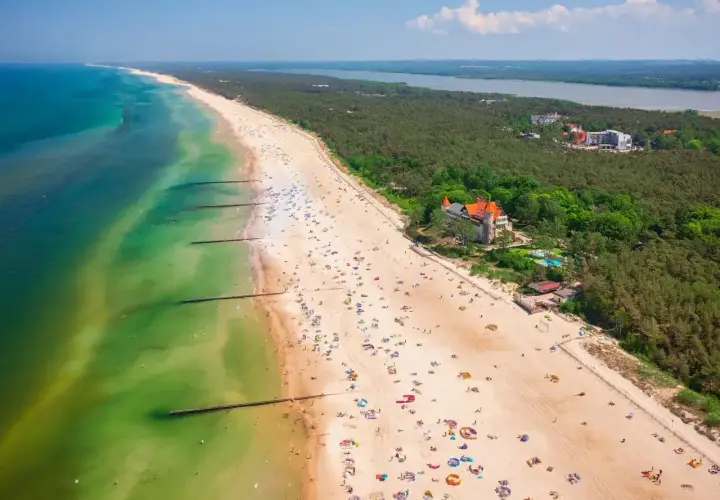 Słoneczna plaża nad Jeziorem Łebsko, pełna wypoczywających ludzi i zielony las. Atrakcje turystyczne w zasięgu wzroku.