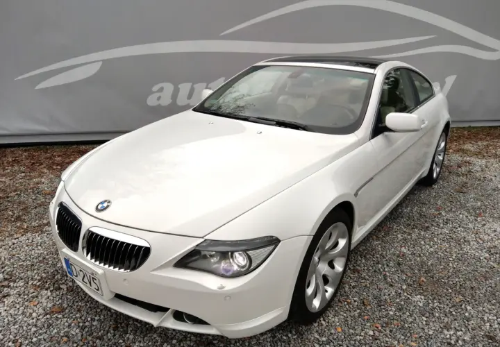 Białe BMW E63 coupe z jasnym wnętrzem, zaparkowane na żwirze, z logo autokomisu w tle.