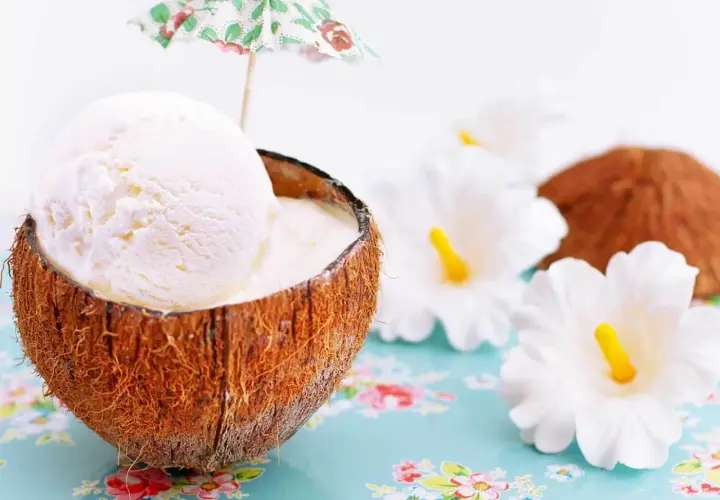 Delicioso helado de coco servido en su cáscara, adornado con una sombrilla tropical y flores blancas.