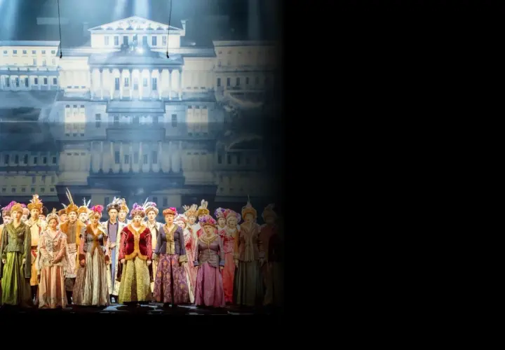 Chór w barwnych strojach historycznych na tle pałacowej scenografii. Sprawdź opera warszawa repertuar!