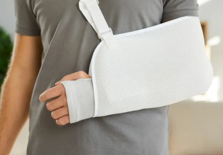 Osoba w szarym t-shircie nosi biały temblak na rękę. Czy trzeba nosić cały czas temblak? To pytanie zadaje sobie wielu po urazie.