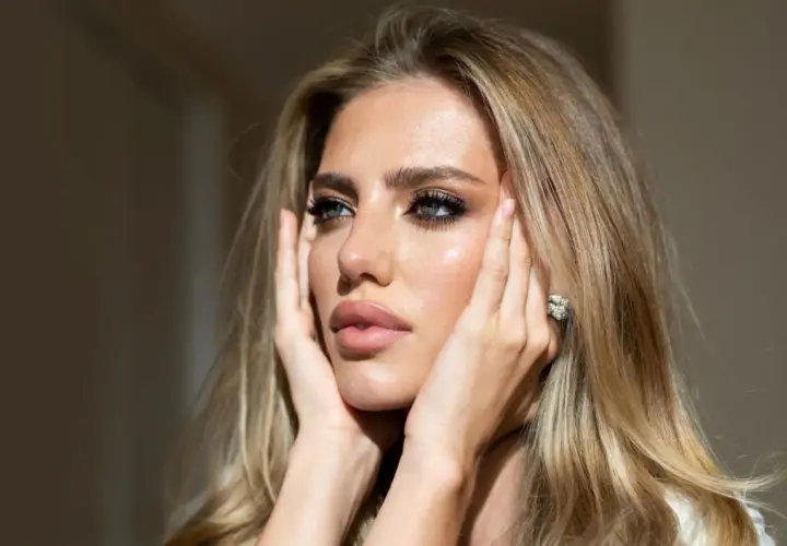 Kobieta z blond włosami i makijażem, dotyka twarzy dłońmi. Widać jej błyszczące usta i oczy. Co pod podkładem? Idealna cera.