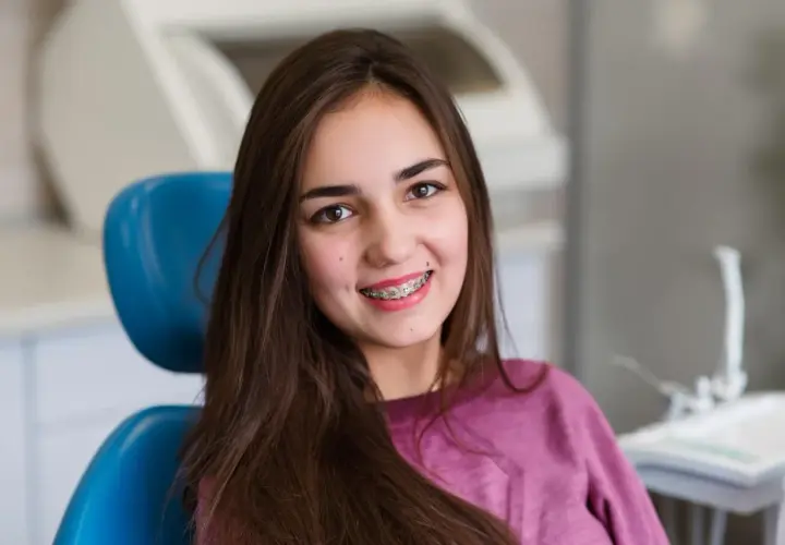 Uśmiechnięta nastolatka z kolorowym aparatem ruchomym na zębach siedzi na niebieskim fotelu dentystycznym.